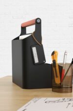 Boîte à outils YouMe noire avec une poignée ergonomique en cuir, posée sur un bureau. À côté, des fournitures comme un cutter, un stylo et des plans d'architecte, idéal pour un environnement de travail professionnel et organisé.