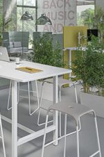 Une table blanche moderne entourée de tabourets élégants, avec des bacs à plantes verdoyants créant une ambiance naturelle dans un bureau lumineux.