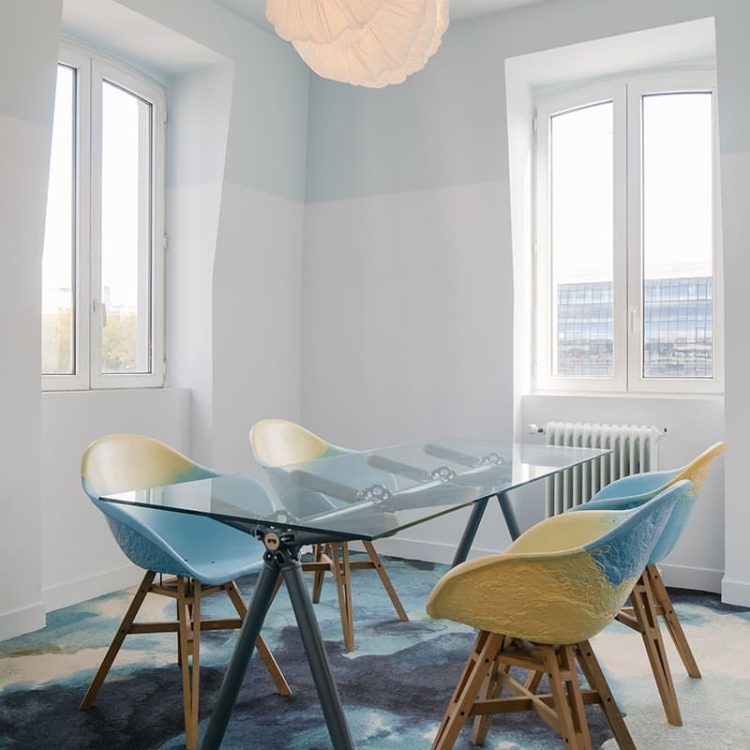 Chaises aux teintes pastel et texturées, en matériaux recyclés, entourant une élégante table en verre, dans un espace lumineux et apaisant.